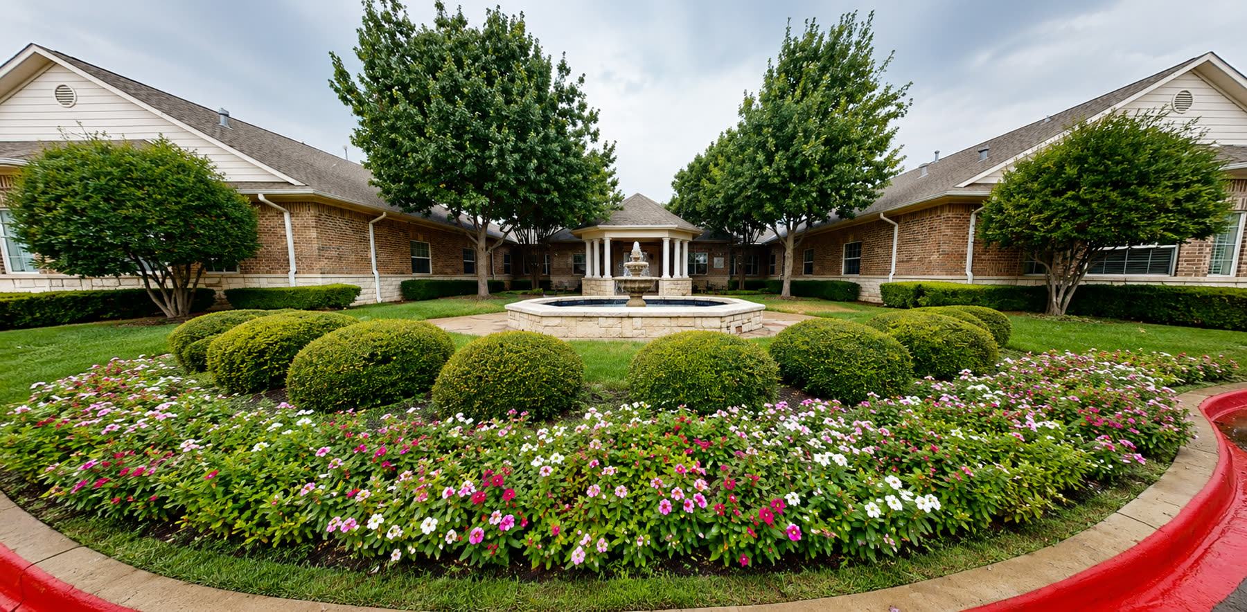 Courtyard fountain surrounded by flower beds and shade trees at Magnolia Memory Care of Flower Mound
